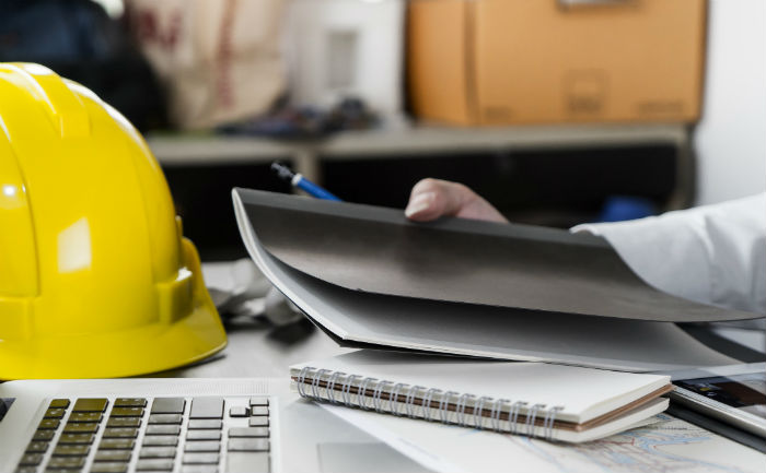 A close-up image of a person holding a black notebook near a laptop and yellow construction hard hat.