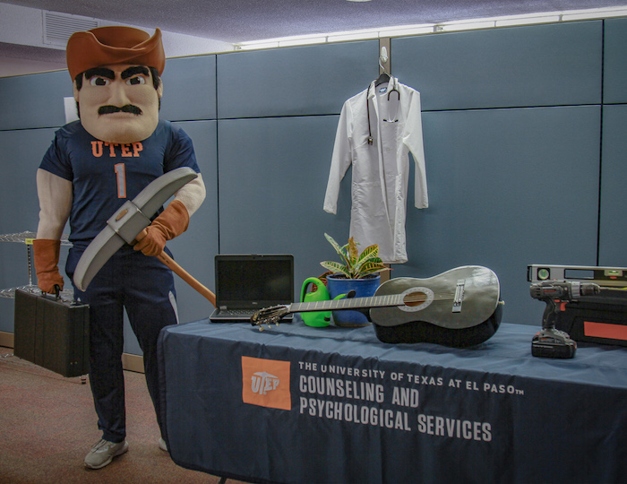 Paydirt Pete standing nets to a Counseling Services table with many different resources like a computer, a plant and pot of water, a guitar, some machinery, and a doctors coat.