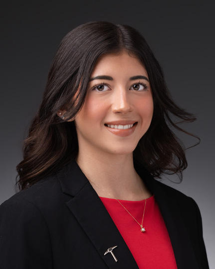 Professional headshot of Elizabeth Hinojosa with shoulder-length dark hair wearing a black blazer, red top, necklace, and pickaxe lapel pin, facing forward against a dark gray studio background.