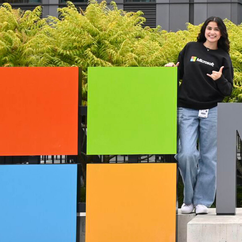 Vianey Martinez wearing a black Microsoft sweatshirt stands beside large colorful Microsoft logo blocks outdoors, posing with a hand gesture in front of greenery and a modern building.