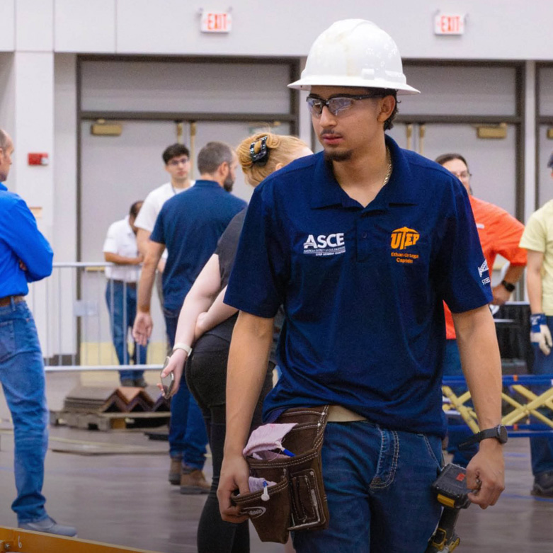 A student wearing a hard hat, safety glasses, a navy UTEP polo shirt with an ASCE patch, jeans, and a tool belt walks through an indoor engineering competition event while carrying a power tool.