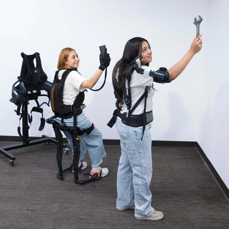 Two individuals in a lab wear full-body exoskeleton systems, with one standing and lifting a wrench overhead while another sits in an exoskeleton device secured to a wheeled support frame and equipment base behind.