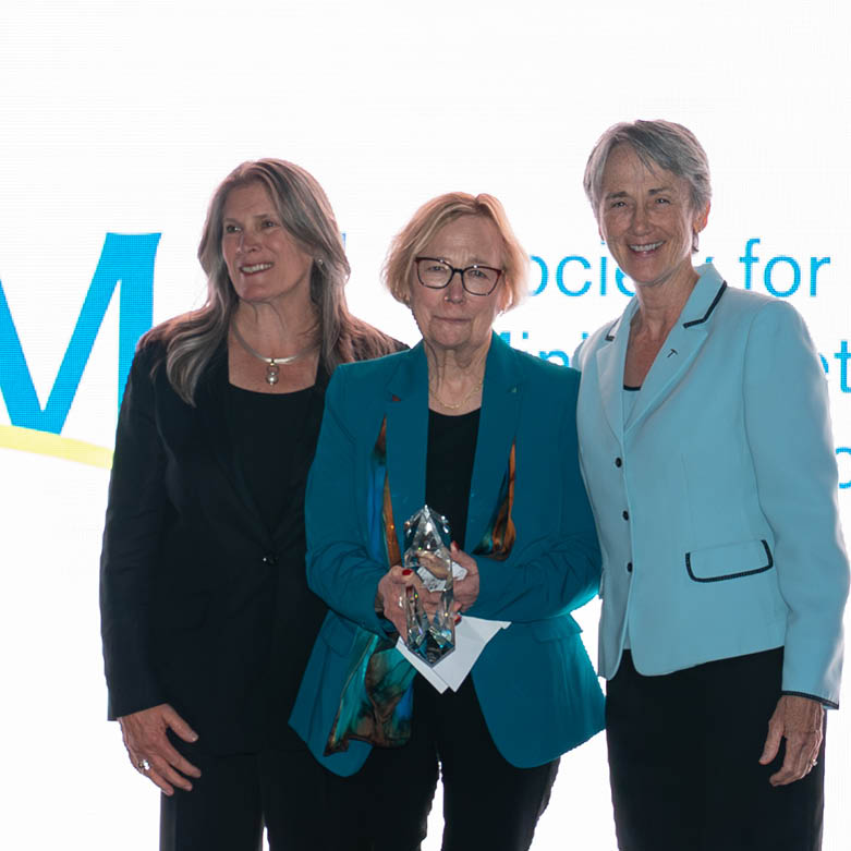 Three individuals on stage at the Miner Business Impact Awards event, with one holding a presidential gold bar award in front of a bright presentation screen.