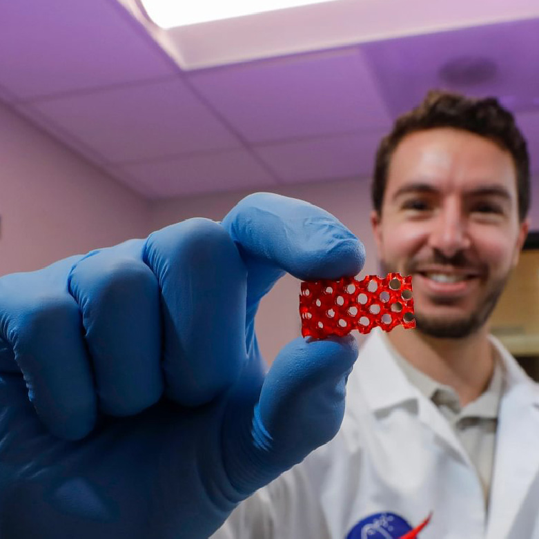A researcher wearing blue laboratory gloves holds a small red 3D-printed battery component with an open lattice design toward the camera, with laboratory equipment and a work area visible in the background.