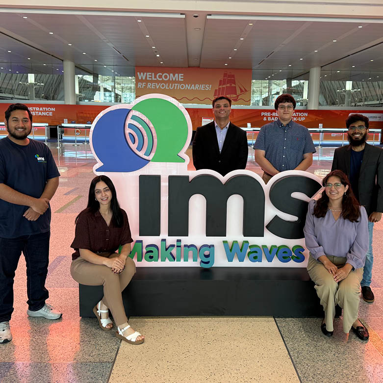 Six students pose around a large ims (lowercase) Making Waves sign in a convention center during the IEEE International Microwave Symposium. Visible text on the sign reads ims Making Waves, and a banner in the background reads Welcome RF Revolutionaries!