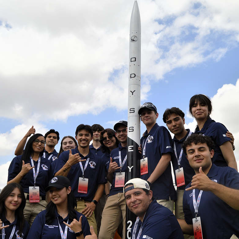A student rocket team wearing matching navy polos and event badges poses beside a tall model rocket labeled ODYSSEY at an outdoor launch site. Team members stand and kneel around the rocket, with grass, a fence, and a cloudy sky visible in the background.