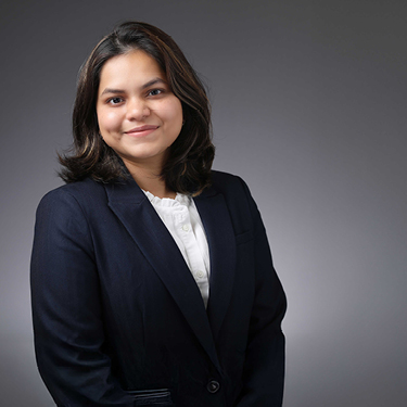 Professional headshot of Anantaa Kotal with shoulder-length dark hair wearing a dark blazer over a white collared blouse, facing forward against a gray studio background.