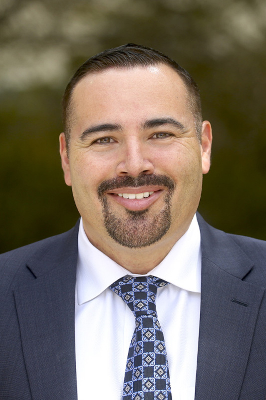 Professional headshot of Rafael Gonzalez wearing a dark suit jacket, white dress shirt, and patterned blue tie, facing forward against a blurred outdoor background.