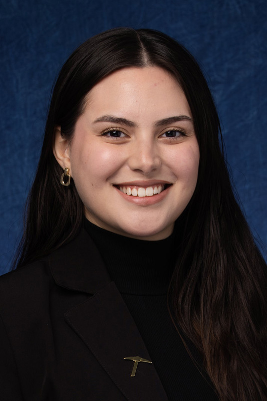 Professional headshot of Alejandra Perez Lopez with long dark hair wearing a black turtleneck and blazer with a pickaxe lapel pin, facing forward against a blue studio background.