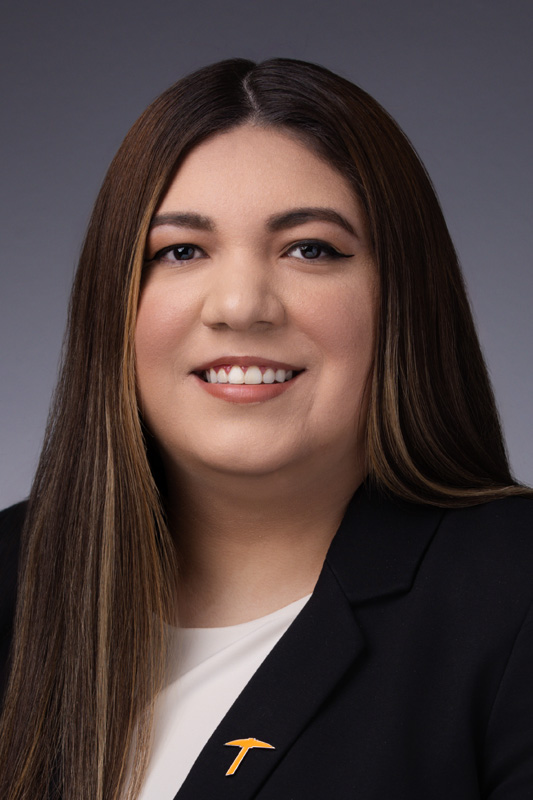 Professional headshot of Alyssa Torres with long straight hair wearing a dark blazer over a white top and an orange pickaxe lapel pin, facing forward against a gray studio background.