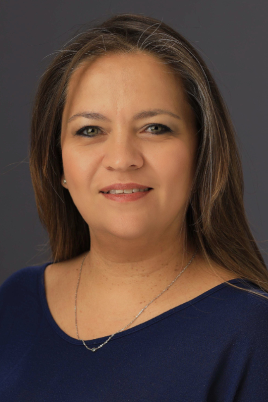 Professional headshot of Ana I. Parham wearing a blue top and a delicate necklace, facing forward against a dark gray studio background.
