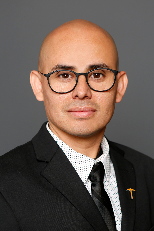 Professional headshot of Eduardo Arreola wearing round glasses, a black suit, a white patterned dress shirt, and a black tie with an orange pickaxe lapel pin, facing forward against a gray studio background.