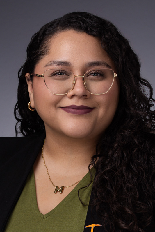 Professional headshot of Marianna Gutierrez wearing glasses, a green blouse, a dark blazer, gold hoop earrings, and a necklace with an M pendant, facing forward against a gray studio background.