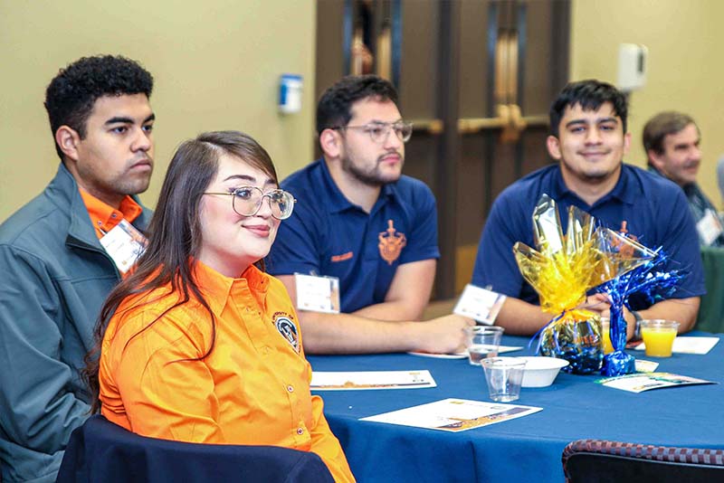 Four attendees seated at a round table listening during an indoor event.