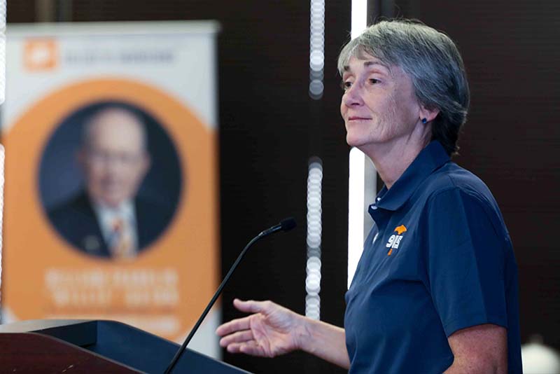 Heather Wilson standing at a podium speaking beside a microphone and a banner in a conference room.