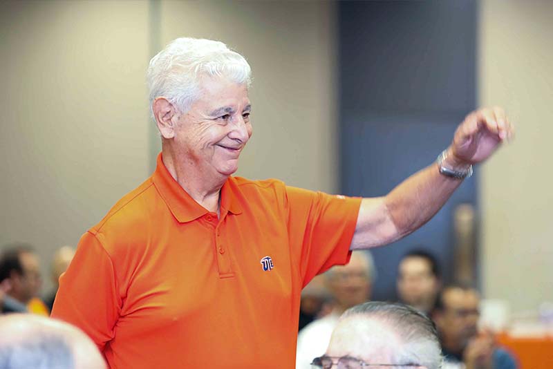 Person standing at a podium speaking beside a microphone and a banner in a conference room.