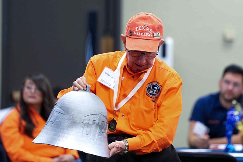 Willie Quinn holding a large metal bell while speaking to attendees seated at tables.
