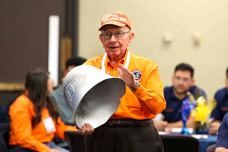Willie Quinn holding a large metal bell during the Loya Engineering 2022 Homecoming breakfast with attendees seated in the background.