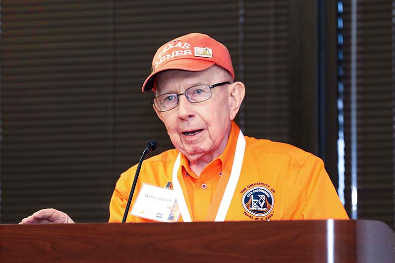 Willie Quinn wearing an orange shirt and cap speaking at a podium with a microphone.