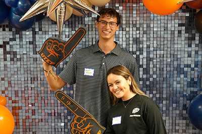 Two attendees pose together in front of a decorative backdrop featuring silver star accents, orange and blue balloons, and a mosaic-style background during a UTEP Engineering homecoming event, holding foam finger props while standing for a commemorative photo.
