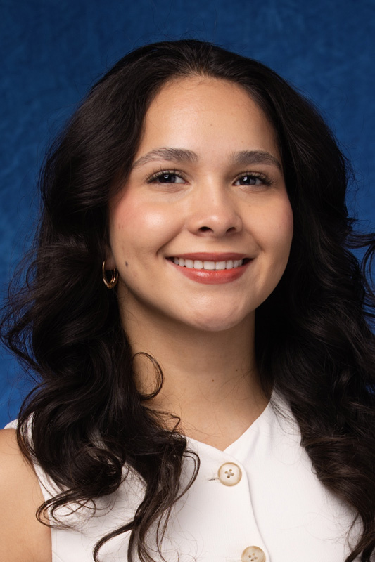 Professional headshot of Adrianna Ruiz with long wavy hair wearing a white sleeveless top and small hoop earrings, facing forward against a blue studio background.