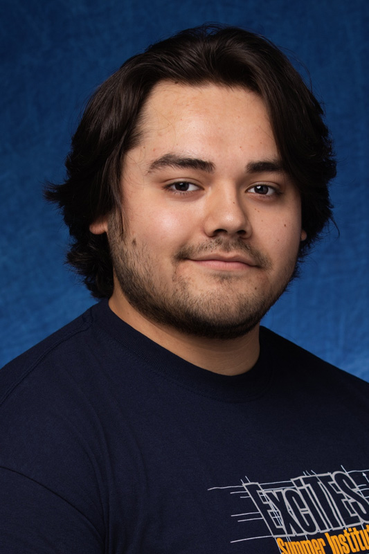 Professional headshot of Arturo Garcia Rivera with medium-length dark hair and facial hair, wearing a dark shirt, facing forward against a blue studio background.