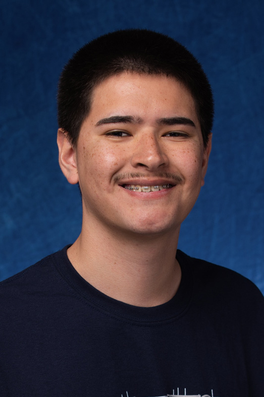 Professional headshot of Diego Del Val wearing a dark shirt, facing forward against a blue studio background with visible braces and short hair.