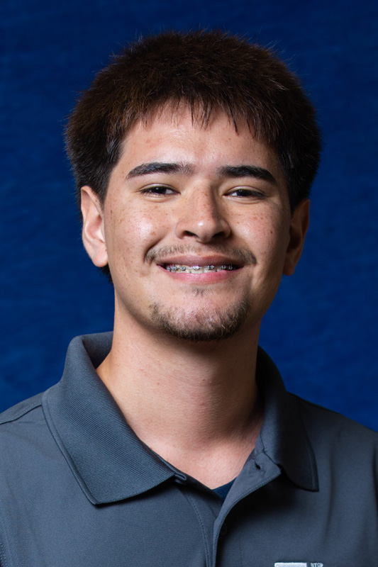 Professional headshot of an individual with short dark hair, braces, and a goatee, wearing a gray UTEP College of Engineering polo shirt and facing forward against a blue studio background.