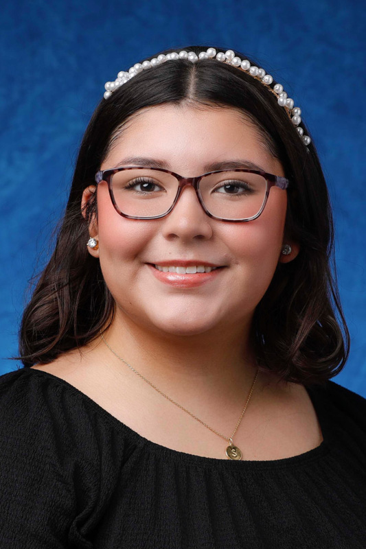 Professional headshot of Sofia Reyes wearing glasses, a pearl headband, stud earrings, and a black top, facing forward against a blue studio background.
