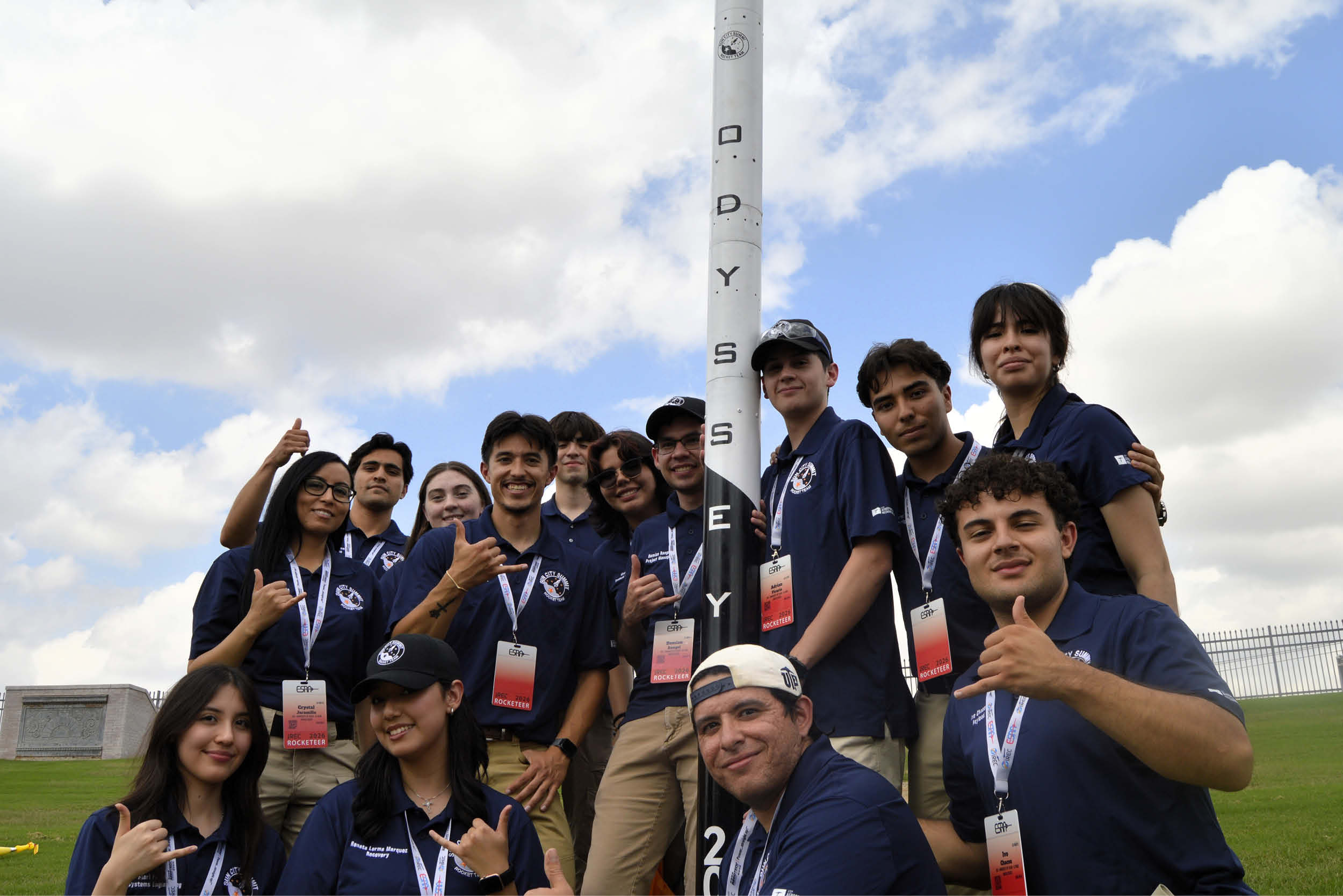 A team of students wearing matching navy polos and event badges gathers around a tall model rocket outdoors, posing for a group photo beneath a cloudy sky. The rocket displays the vertical text ODYSSEY, and several team members make hand gestures toward the camera.