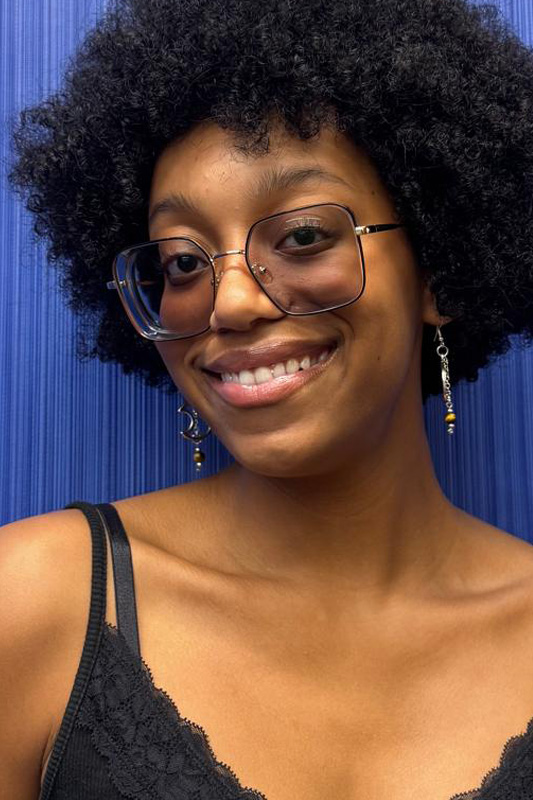 Professional portrait of Ava Napier wearing large rectangular glasses, dangling earrings, and a black sleeveless top, posed against a textured blue background.