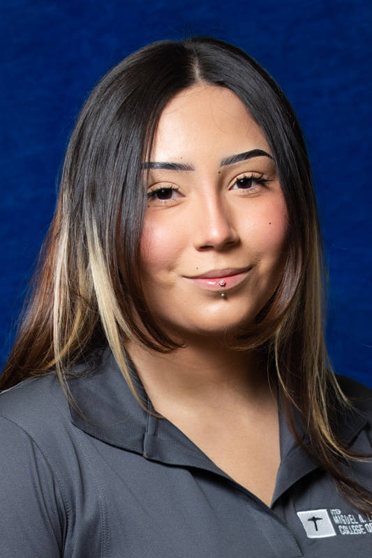 Professional headshot of Tzetzaith Rivero with long dark hair featuring lighter highlights, wearing a gray UTEP College of Engineering polo shirt and a lip piercing, facing forward against a blue studio background.