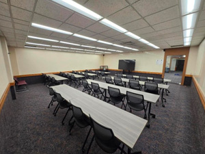 Classroom with rows of tables and chairs arranged for instruction, featuring a wall-mounted display screen, carpeted flooring, and overhead fluorescent lighting.
