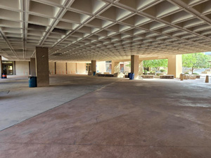 Covered outdoor breezeway with a geometric coffered ceiling, wide concrete walkways, and support columns, providing an open pedestrian space between campus buildings.