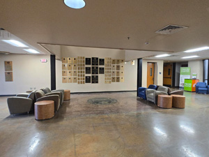 Open lobby area with polished concrete floors, lounge seating, colorful chairs, and a wall displaying plaques and awards, creating a welcoming gathering space within a campus building.