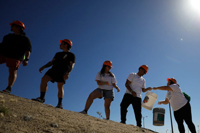 Participants wearing orange hard hats stand along a sloped, rocky hillside, passing buckets between each other during an outdoor activity. The scene is backlit by a bright sun in a clear blue sky, creating strong shadows and silhouettes against the terrain.