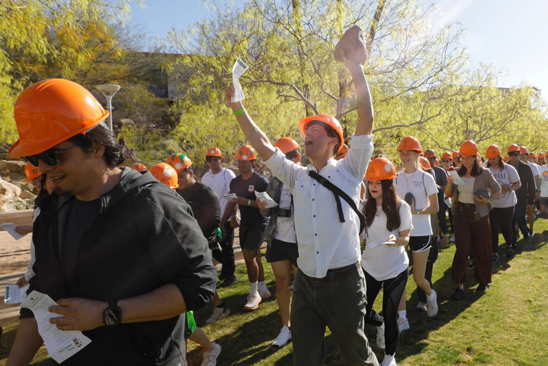 Participants wearing orange hard hats walk together across a grassy campus area, each holding papers as part of an organized outdoor activity. One participant in the center raises both arms while others follow in a line, with trees and campus buildings visible in the background under bright sunlight.