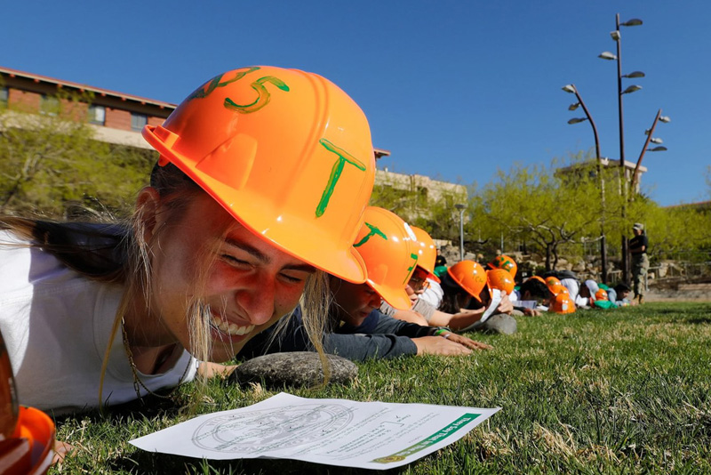 Student participants wearing orange hard hats lie face down on a grassy field, closely examining a printed sheet as part of a hands-on outdoor activity. Additional participants in helmets are lined up behind them near campus buildings under a clear blue sky.
