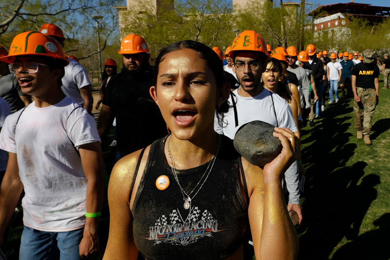 Participants wearing orange hard hats walk in a line across a grassy campus area, with one individual in the foreground carrying a large rock on their shoulder. Several others follow closely behind, some marked with colored powder, as campus buildings and trees appear in the background under bright sunlight.