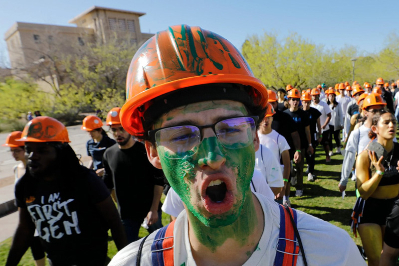 Close-up of a participant wearing an orange hard hat with green paint markings and glasses, with green paint covering part of the face, standing at the front of a line of participants during an outdoor campus activity. A group of similarly dressed participants follows behind on a grassy area with trees and campus buildings visible under bright daylight.