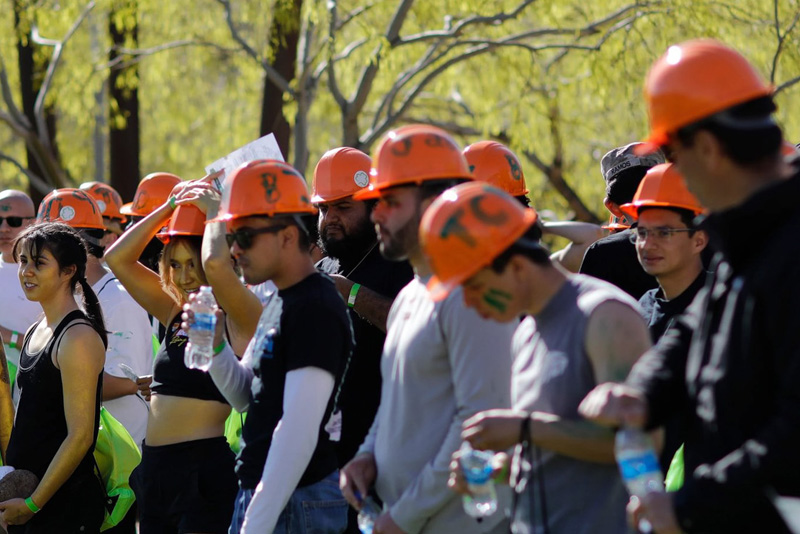 Participants wearing orange hard hats stand together on a grassy campus area, holding water bottles and papers while taking a break during an outdoor activity. Trees with green foliage and campus buildings appear in the background under bright sunlight.