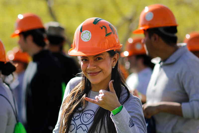 Participant wearing an orange hard hat marked with green paint faces the camera and makes a hand gesture, standing among a group of similarly dressed participants during an outdoor campus activity. Other participants in hard hats appear in the background on a grassy area with trees under bright daylight.