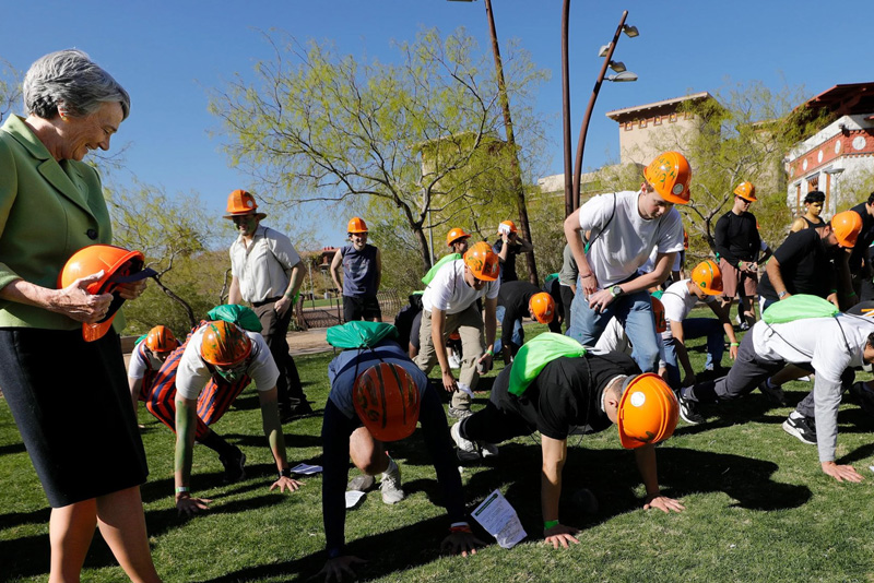 Participants wearing orange hard hats perform group exercises such as push-ups on a grassy campus lawn while holding or keeping track of papers. President Wilson stands to the side holding a helmet and observing, with trees and campus buildings visible in the background under clear daylight.