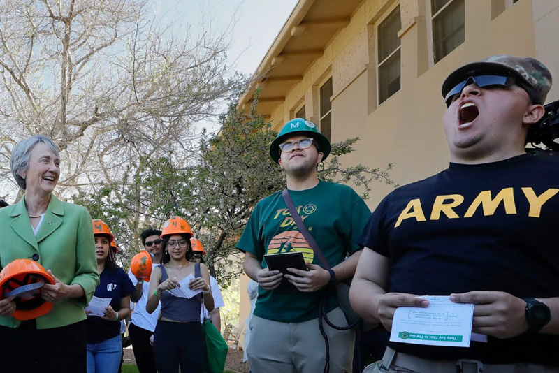 A group of participants wearing orange hard hats gathers outdoors, holding papers and appearing to chant or recite together during a campus activity. Some participants wear themed shirts such as one reading ARMY, while trees and a building form the background in bright daylight.