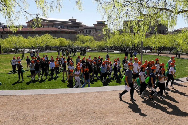 Large group of participants wearing orange hard hats walks together across a grassy campus area, many holding papers during an organized outdoor activity. Trees with fresh leaves and university buildings line the background under a clear sky.