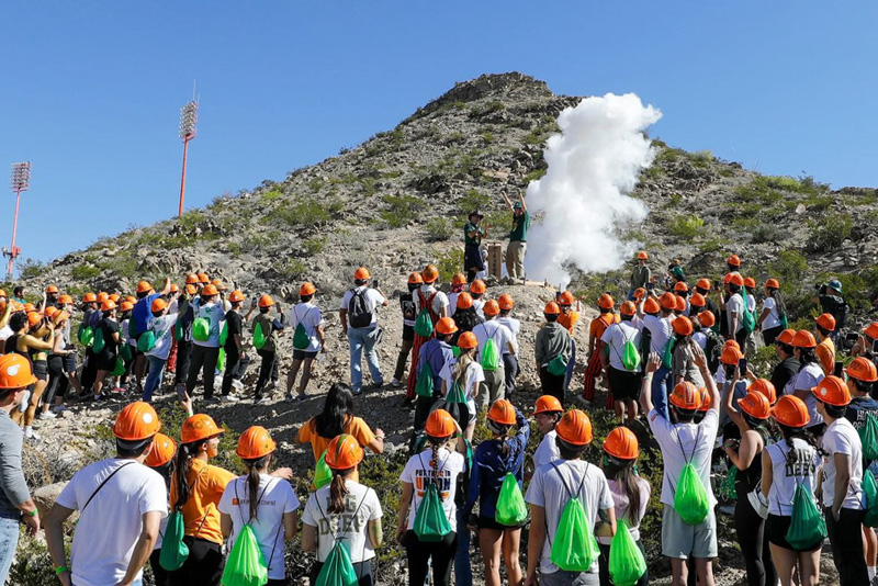 A large group of participants wearing orange hard hats gathers on a rocky hillside, facing a small group at the top where smoke rises during an outdoor campus activity. Many participants carry green bags, with a mountain and clear blue sky forming the background.