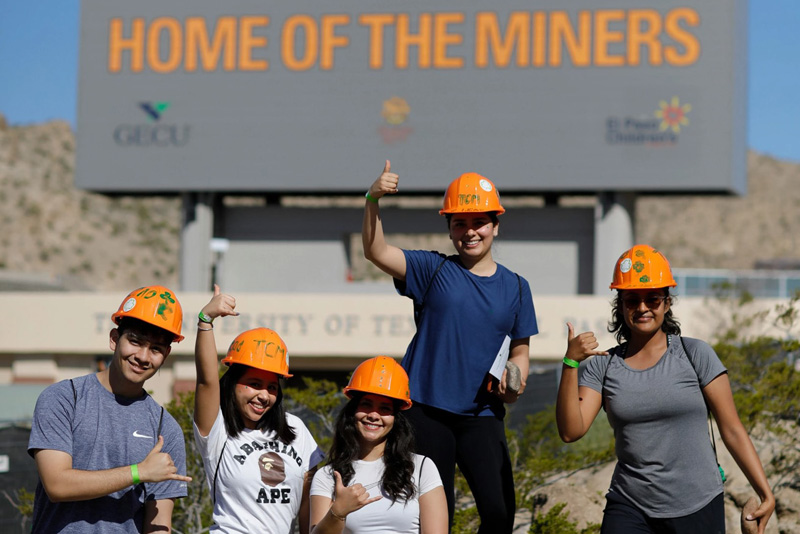 Five participants wearing orange hard hats pose together outdoors, each making a thumbs-up gesture while standing in front of a large sign that reads HOME OF THE MINERS. The group stands on a rocky area with campus buildings and mountains visible in the background under bright sunlight.