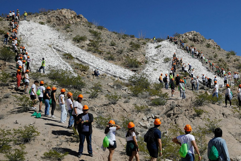 A large group of participants wearing orange hard hats spreads across a rocky hillside, forming lines as they hike up paths of light-colored rock. Some individuals pause along the slope while others continue climbing, with sparse desert vegetation and a clear blue sky in the background.