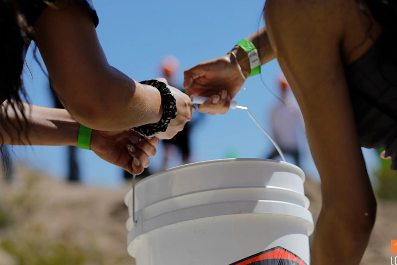Close-up of participants’ hands passing a white bucket along a line during an outdoor activity, with green wristbands visible on their wrists. The background shows a sunlit hillside with blurred figures wearing orange hard hats against a clear blue sky.
