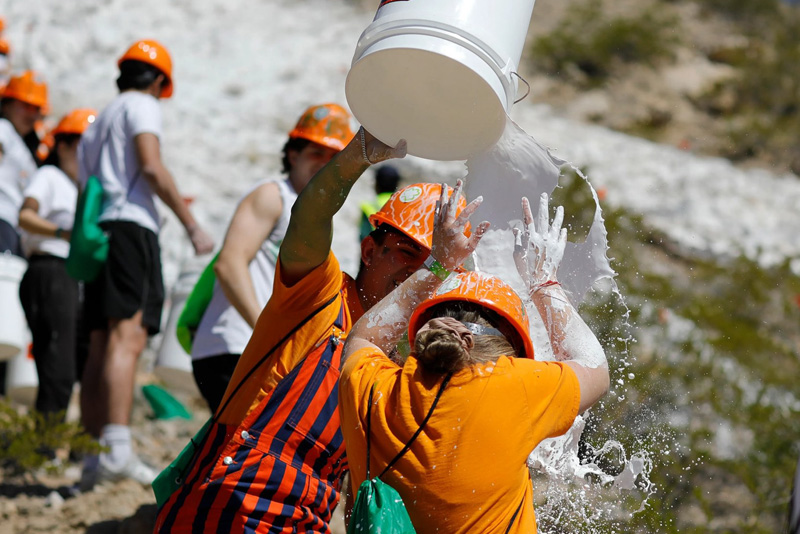 Participant wearing an orange hard hat pours a bucket of white liquid over another participant’s head on a rocky hillside, splashing across both individuals during an outdoor activity. Other helmeted participants stand nearby, with a large white-painted section of the mountain visible in the background under bright sunlight.